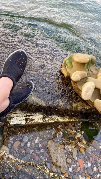 Feet and legs of a woman by the sea and on the edge of a rock. The woman has sea shoes on her feet
