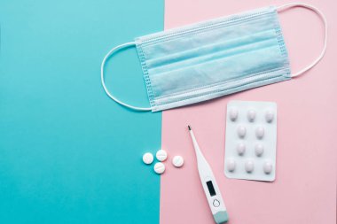 Koronavirüs enfeksiyonuna karşı koruma: tek kullanımlık mavi maske, beyaz haplar, açık pembe ve turkuaz arka planda elektronik termometre.