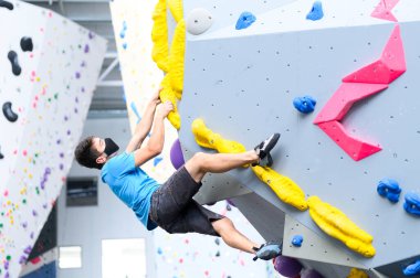 COVID-19 salgın maskesi takan bir adam sosyal mesafeyi takip ederek tırmanma salonundaki kaya duvarına tırmanıyor.