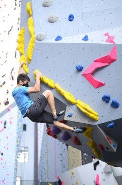 COVID-19 salgın maskesi takan bir adam tırmanma salonundaki kaya duvarına tırmanıyor. Egzersiz için sosyal mesafe kurallarına uyuyor.