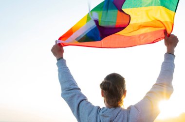 Elinde neşeli bir gökkuşağı bayrağı tutan bir adamın silueti. Rüzgarda dalgalanan tropik bir plajda altın güneş ışığıyla.