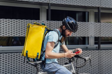 Bisikletle yiyecek getiren sakallı genç kurye akıllı bir telefonla siparişi kontrol ediyor. Teslimat hizmeti kavramı.