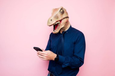 Dinozor maskeli adam akıllı telefon kullanıyor. Pembe arka plan.