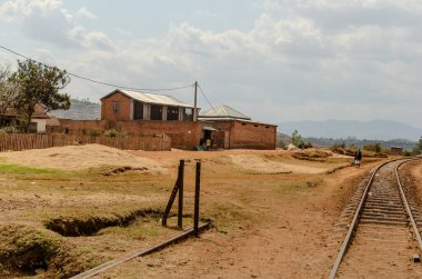 Madagaskar 'da sakin bir kırsal alan. Kuru ve toprak arazide kıvrılan tren rayları var. Görüntü, ahşap bir çitle çevrili metal çatılı küçük bir tuğla bina yakalıyor. Sahne yumuşak, doğal bir banyo.