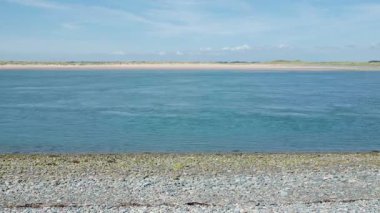 Deniz akıntısı, Morfa Dinas 'taki çakıl taşı kıyısı ile Newborough, Menai Boğazı, Galler' deki kumlu plaj arasındaki dar sesten geçiyor. Açık mavi gökyüzünün altında, bir yaz günü..