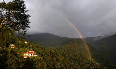 Canlı bir gökkuşağı, yoğun yeşil dağların üzerinde zarif bir şekilde yamaçlarda altın ışıkta parlayan evlerle parıldıyor. Huzurlu bir sahne doğanın güzelliğini, uyumunu ve yağmur sonrası tazeliğini yansıtıyor..