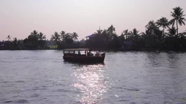 9 saniyelik bir HD videoda Kerala durgun sularında süzülen yolcu botu, siluetli palmiye ağaçları ve sakin kıyı şeridi boyunca uzanan uzak evler görülüyor..