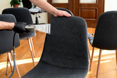 Eldiven takan bir kişi gri döşemeli sandalyeyi çağdaş bir ortamda yenilemek için buhar temizleyicisi kullanır. Sıcak ahşap zemin ve mobilyalar rahat bir atmosfer yaratır..