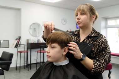Şık bir kuaförde, yetenekli bir stilist genç bir çocuğun saçlarını titizlikle keser, taze bir görüntünün zevkini yakalar. Atmosfer canlı, renk ve yaratıcılıkla dolu..