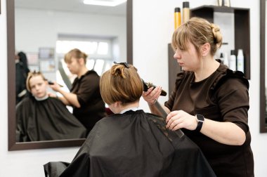 Yetenekli bir kuaför, çağdaş bir kuaförde müşterinin saçını dikkatlice keser. Parlak alanda aynalar ve çeşitli saç ürünleri var. Rahat bir atmosfer yaratıyorlar..