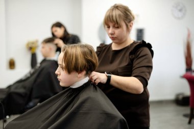 Bir çocuk pelerinle kaplı bir salonda otururken bir berber saçını ustalıkla şekillendiriyor. Şık bir ortam öğleden sonra saç kesiminin rahatlatıcı atmosferini arttırır..