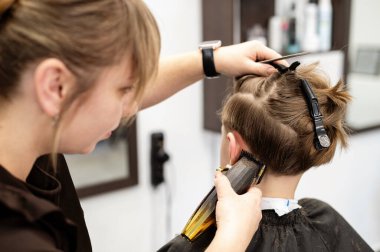 Yetenekli bir stilist çağdaş bir kuaförde bir çocuğun saçını dikkatlice keser. Çocuk sakince oturup koruyucu bir pelerin giyerken kuaför makas kullanıyor..