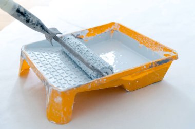 Bir silindir, içi taze beyaz boyayla dolu turuncu bir tepsiye batırılır. Temiz, beyaz arka plan, ev geliştirme veya sanatsal yenileme sürecini vurguluyor.