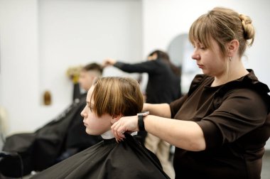 Bir stilist saçını dikkatlice keserken küçük bir çocuk kuaför koltuğunda oturur. Çevre aydınlık ve davetkar, diğer müşteriler yakınlarda..