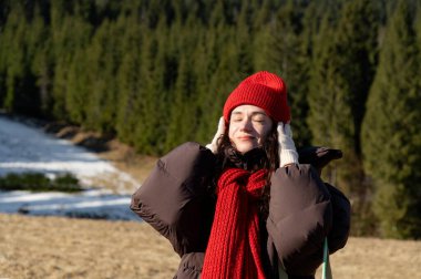 Sakin bir orman ortamında, bir kadın gözleri kapalı, kışın serinliğini kucaklarken canlı kırmızı bir şapka ve eşarp takıyor. Huzurlu ve mutlu görünüyor..