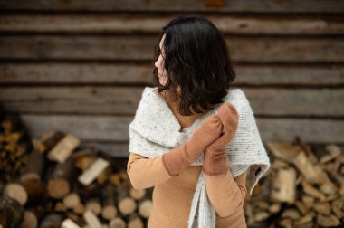 A young woman stands outside in winter attire, wearing a soft scarf and gloves while near a woodshed. She enjoys the serene atmosphere, embodying wellness and mindfulness. clipart