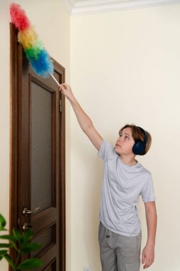 Bir genç, renkli bir toz beziyle kapı çerçevesinin tozunu alırken ev ödevine odaklanır. Güneş ışığı odayı aydınlatır, çalışmak için davetkar bir atmosfer yaratır..