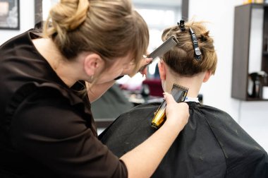 Yetenekli bir kuaför, çağdaş bir kuaför ortamında makas ve makas kullanarak genç bir müşterinin saçını dikkatlice keser. Müşteri oturmuş ve pelerinle kaplanmış..