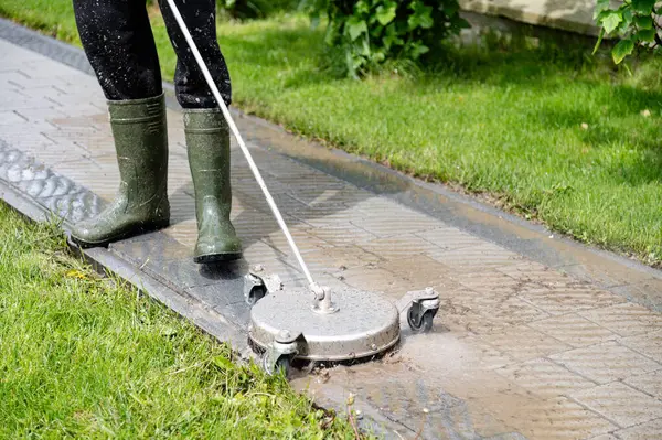 Yeşil bir çimenlikte duran bir insan asfalt bir yolda yüzey temizleyicisini çalıştırıyor. Temizlik işlemi su püskürtüyor ve toprağı temizliyor. Böylece etkin bir dış bakım aktivitesi ortaya çıkıyor. Profesyonel kaldırım taşı temizliği.