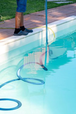 Güneşli bir öğleden sonra evlerin arka bahçesinde havuzu elektrikli hortumla temizleyen kişi.