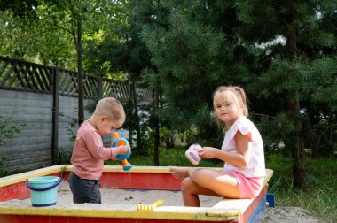 A father and his two children play in a colorful sandbox in their backyard, surrounded by greenery. The kids are engaged and happy, enjoying a beautiful day outdoors.