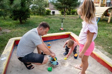 A father plays with his children in a sandbox, filled with colorful toys. They dig and build, laughing together in a joyful park setting during a bright afternoon.