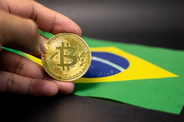 Arka planda Brezilya bayrağı olan Bitcoin 'i tutan bir el. Dijital para birimini ve engelleme teknolojisinin küresel ve yerel ekonomilerle ilişkisini sembolize ediyor.