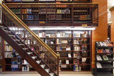 Ahşap merdivenleri ve geniş kitaplıkları olan iki katlı modern bir kütüphane. Mekanda sıcak ışıklandırma, kitap depolama seviyeleri ve metal parmaklıklar ve düzenli okuma materyalleri ile çağdaş mimari tasarım yer alıyor..