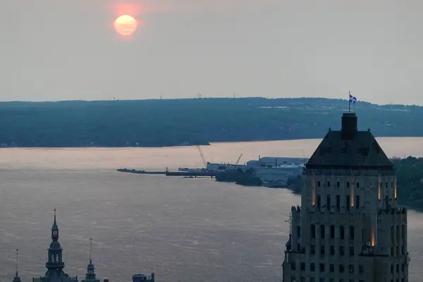 Gölün ya da büyük bir su kütlesinin üzerinde güzel bir gün doğumu manzarası. Sabah güneşi ufukta beliriyor siluetli kıyı şeridi ve binalar ön planda. Sakin su yüzeyine yansıyan ılık turuncu ve pembe gökyüzü renkleriyle huzurlu şafak atmosferi. 