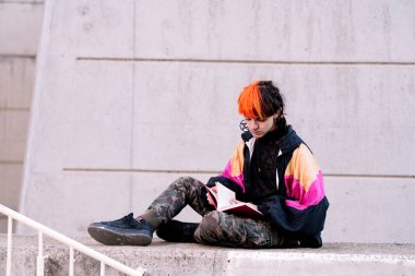 Turuncu ve siyah saçlı çocuk sokakta kitap okuyor..