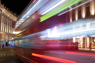 Londra, Birleşik Krallık 6 Ekim 2013: Regent Caddesi 'ndeki renkli ışık yollarıyla hareket eden kırmızı çift katlı bir otobüsün uzun pozlu fotoğrafı. Arka planda klasik binalar, İngiliz bayrağı ve parlak şehir ışıkları görülüyor..