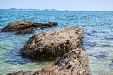 Kıyı manzarası geniş, engebeli kayaları kısmen turkuaz sulara batırır. Uzak dağlar güneşli bir günde huzurlu bir zemin oluştururken kayalara doğru hafif dalgalar süzülür..