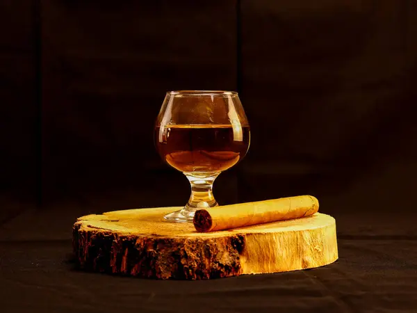 Bir bardak viski ve puro, kasvetli arka planda, ahşap bir dilimin üzerine konmuş. Minimalist ve zarif bir düzen, lüks için mükemmel, erkeksi yaşam tarzı, ve kutlama teması