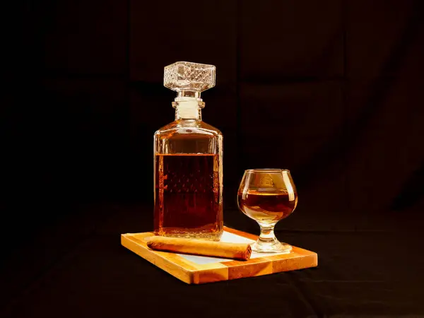 Zarif bir viski sürahisi ve koyu bir arka planda duran yuvarlanmış puroyla ahşap bir tepside servis edilen cam. Lüks, erkeksi yaşam tarzı, kutlama ve kaliteli alkol markası teması için mükemmel.