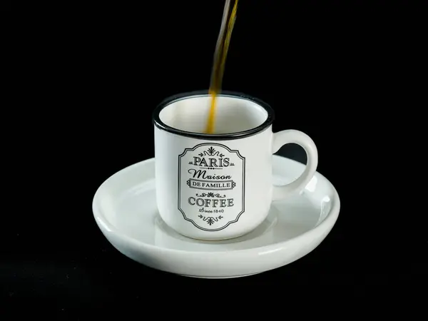 Beyaz seramik bir fincana koyu kahve dökülüyor, Paris yapımı klasik bir tasarım, siyah kumaş arka planına uygun bir fincan çay tabağına yerleştiriliyor. Kafein, kahvaltı, kahve kültürü ve sabah ayinleriyle ilgili kavramlar için mükemmel.