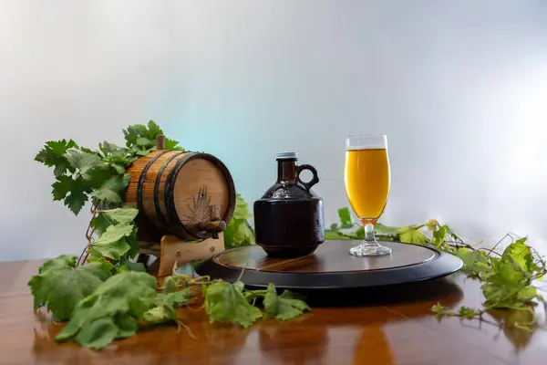 Bir bardak altın zanaat birası koyu bir homurtunun yanında köpüklü bir kafa ve yeşil hop yapraklarıyla çevrili dekoratif ahşap bir fıçı..