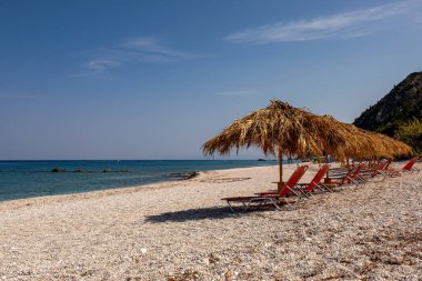 Güzel boş Akdeniz sahilleri sıralar halinde trav şemsiyeleri ve kızıl güneş şezlongları, berrak mavi deniz, çakıllı sahil ve parlak yaz gökyüzü.