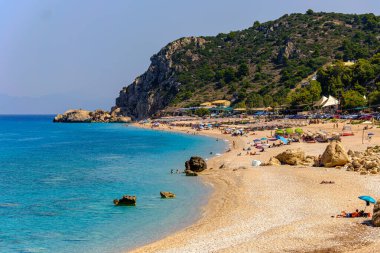 Yunanistan 'ın Lefkada Adası' ndaki Kathisma Sahili 'nin geniş panoramik manzarası. Kumsal güneşlenenlerle, renkli şemsiyelerle ve turkuaz İyon Denizi dalgalarıyla dolu. Tepeler ve kayalar sahneyi çevreleyerek çarpıcı bir tatil atmosferi yaratıyor..