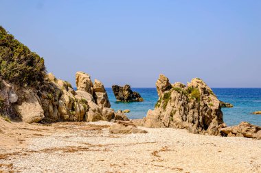 Denizin sakin, turkuaz sularına ve berrak mavi gökyüzüne karşı kurulmuş altın kum ve çakıl taşlarından yükselen çarpıcı doğal kaya oluşumlarının yer aldığı huzurlu plaj sahnesi. Doğa ve seyahat için ideal..