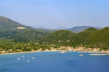 Geniş bir Vassiliki Körfezi manzarası, Lefkada, demirli yelkenli tekneleri, büyüleyici bir köy, ve berrak bir gökyüzü altında yemyeşil dağlar. Seyahat, yelkencilik ve sakin Akdeniz temaları için mükemmel..