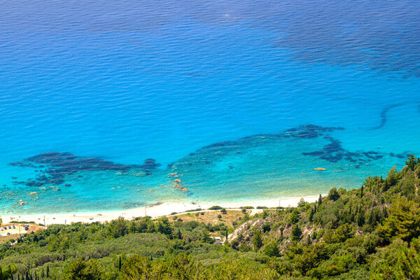 Stunning aerial view of Lefkada's pristine coast. Lush green hills meet a white beach and crystal-clear turquoise waters. A serene natural paradise with vibrant colors and visible road access.
