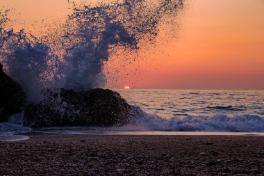 Akdeniz 'de gün batımının canlı turuncu ve pembe renkleriyle aydınlanan Lefkada, Megali Petra Sahili' ndeki bir kayaya güçlü bir dalga çarpar. Dinamik hareket, sakin alacakaranlıkla buluşur..