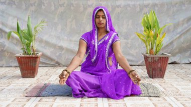 Uzun boylu, düşünceli, genç Hintli kadın Mudra jesti yapıyor. Evde rahat bir koltukta lotus pozisyonunda oturuyor. Huzurlu milenyum kızı derin meditasyon yapıyor, yoga egzersizleri yapıyor..