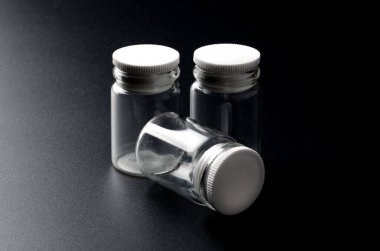 Stüdyo aydınlatmasında koyu dokulu bir yüzeye yerleştirilmiş beyaz plastik kapaklı üç küçük şeffaf cam kavanoz ambalaj, depolama veya ilaç kavramları için idealdir.