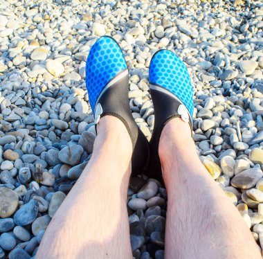 Bakış açısına göre mavi su ayakkabılı bacaklar güneşli çakıl taşlı kumsalda dinleniyor.