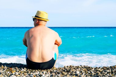 Plaj şapkalı bir adam çakıl taşlarında oturuyor ve sakin denize bakıyor.