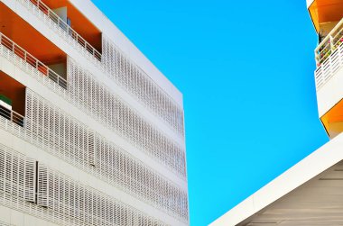 Parlak mavi gökyüzünün altında balkonları olan beyaz ve turuncu binalar modern mimari çizgiler gösteriyor.