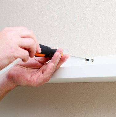 Adam beyaz rafı tornavida kullanarak duvara yapıştırıyor, ellere ve yerleştirmeye konsantre oluyor..