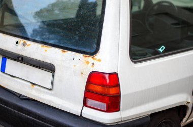 Eski beyaz bir arabanın paslı arka köşesi. Arka lambası ve boş plakası var.
