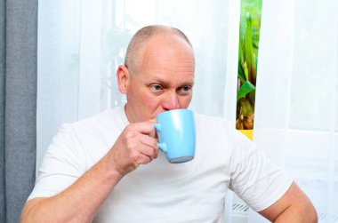 Beyaz tişörtlü, elinde mavi bardakla pencereden dışarı bakan olgun kel adam.